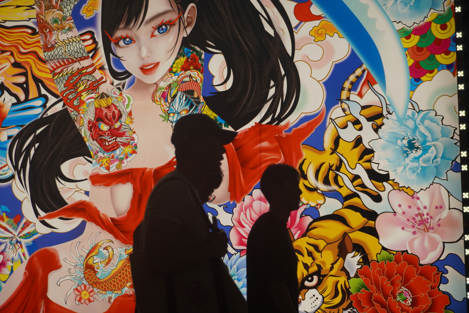 Silhouette of group of young Japanese walking past a bright animated display of a sexualized female character.